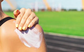 Fitness woman is applying sunscreen on her shoulder before training at the stadium. Protect your skin during sport activity.